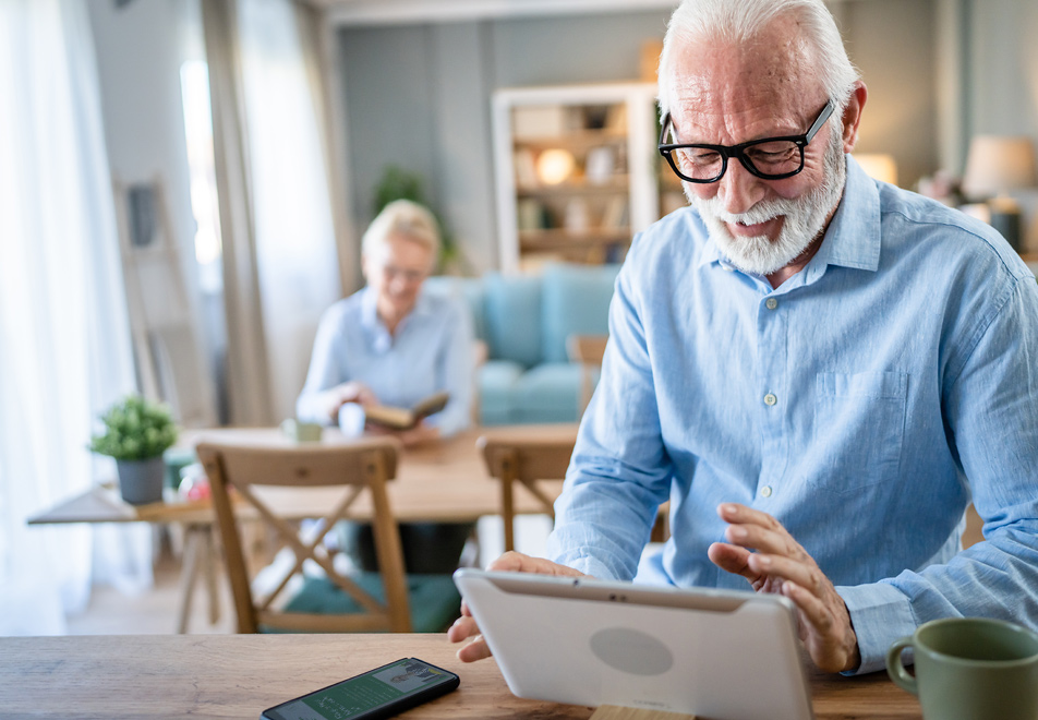 Älterer Mann am Tablet, Frau im Hintergrund